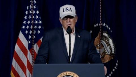 US President Donald Trump stands at a podium wearing a white cap with 'USA' written on it