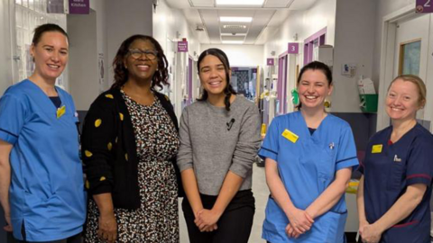 Jasmine Page is pictured smiling on a hospital ward, she is standing in between four nurses. She wears a grey jumper and black trousers with black trainers. Three nurses are wearing blue nurses uniform with their hair away from their faces while another nurse wears a black and white floral dress, black and yellow cardigan and glasses.