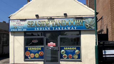 A general view of Seaside Tandoori. It is a one storey building with a pitched roof, and two windows at the side of the main door. There is a sign above the door and a sign with pictures of dishes at either side of the door.