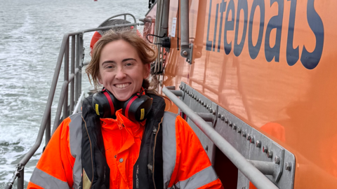 A smiling Hatty McKay stands on the deck of an orange lifeboat. She is wearing an orange hi-viz jacket and ear defenders around her neck.