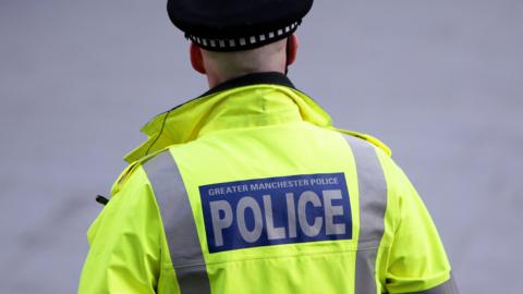 The back of a Greater Manchester policeman in yellow hi-vis jacket with GMP branding and wearing a police cap