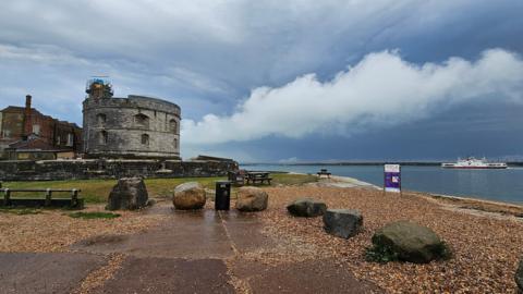 A small stone beach surrounded by boulders sits in front of a body of water containing a white boat in the distance at Calshot. In the centre of the shot is Calshot Castle, a small round grey stone fort.