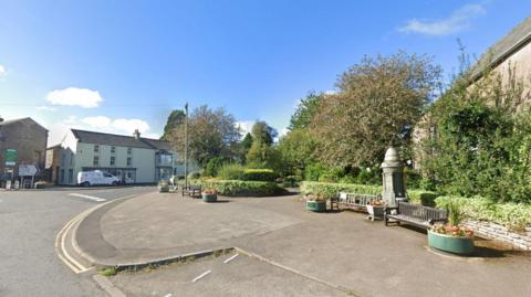 The entrance to the Silver Street memorial garden at Kirkby Stephen. There are two wooden benches on the pavement along with a memorial sculpture. There are also four flower pots spaced along the street. In the background are trees and bushes.