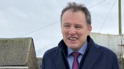 Shows a man smiling in a blue shirt, purple, navy blue overcoat and standing in front of telegraph pole with cloudy sky in the background