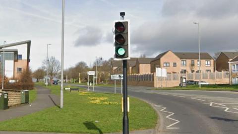 A road curves away into the distance. There is a patch of grass and then buildings on the left and buildings on the right. A traffic light on green in the foreground, white road signs in the distance and street lamps on either side of road.