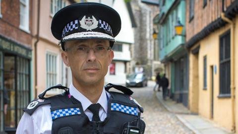 Chief Constable Paul Sanford standing in uniform on a cobbled street in Norwich lined with Tudor buildings.