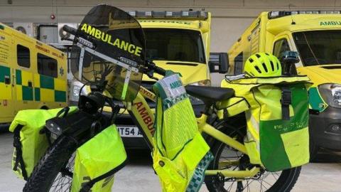 A bright yellow emergency response bicycle equipped for ambulance duties. It has large pannier bags on both the front and rear sides, also in fluorescent yellow with reflective strips for safety. The front panniers are mounted on either side of the wheel, and the rear panniers appear to carry medical supplies. There are ambulances in the background.