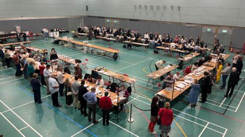A sports hall full of people standing one side of long tables opposite people sitting down with papers spread out in front of them.