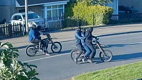 Two masked people ride one motorbike, while three masked people ride another motorbike nearby. All five of them are wearing dark jackets, jeans and balaclavas. 