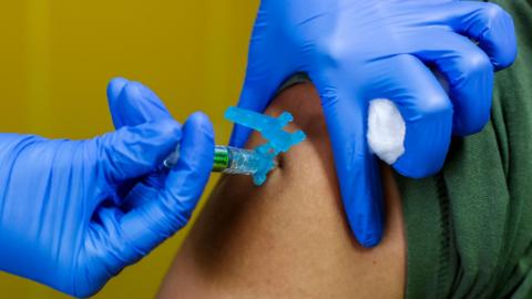 A close up of a persons arm receiving a vaccine. Another person is wearing blue gloves and administering the shot