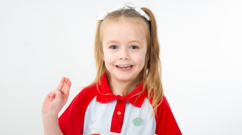 Merch fach mewn crys polo coch a gwyn. Mae ganddi rhubanau gwyn yn ei gwallt melyn.