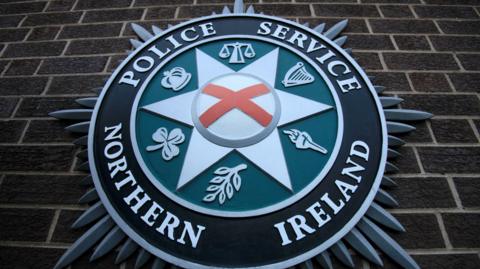A close up of the PSNI emblem on a brick wall. It consists of a star shape in the centre, with the scales of justice, a harp, a torch, a leaf, a shamrock, and crown.