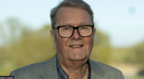 A man with glasses on and a green bush behind. 