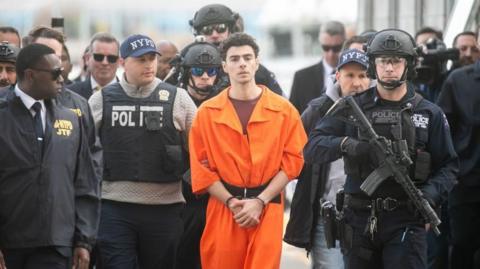   Luigi Mangione is in an orange prison jumpsuit which he is walked while handciffed extradited alongside several armed officers. They are outside having just gotten off a helicopter in New York. His face has no expression.