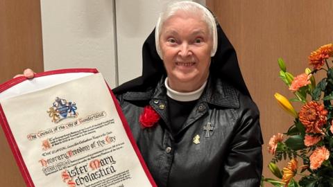 Sister Mary Scholastic stands wearing a black leather jacket holding her Freedom of the City of Sunderland scroll, next to a bright orange flowers at Sunderland City Hall. 