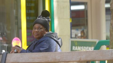 A woman sits on a bench in a town centre looking at camera.