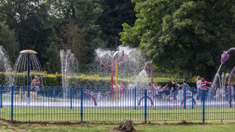 Several water fountains are spraying upward and outward from colorful structures. Children and a few adults are gathered inside the splash area, enjoying the water.
