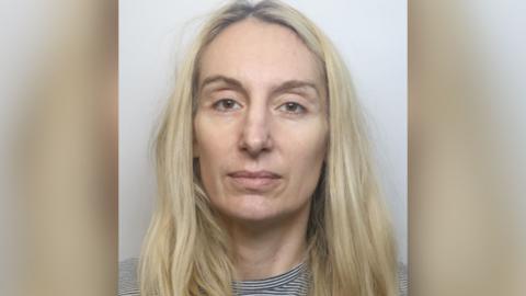 A custody shot of Gemma Kingsley. She has long blonde hair and is wearing a black and white striped top. She is looking at the camera with a blank expression.