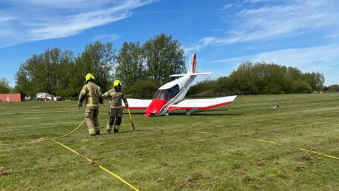 Two fire officers in brown and yellow uniform stand in front of a small plane which is painted white and red. The plane is facing at an angle down into the field, with its nose crumpled. There are trees behind and blue sky.