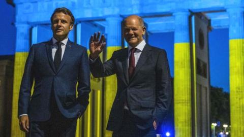 German Chancellor Olaf Scholz and French President Emmanuel Macron (L) wave at the Brandenburg Gate illuminated in the colors of the on May 9, 2022