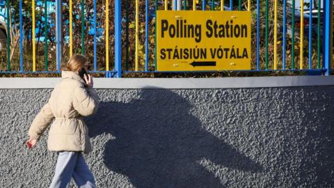 A woman in a beige coat and grey trousers walks past a yellow sign on a fence that says 'Polling Station' in English and Irish.