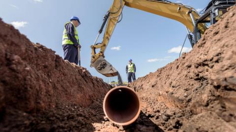 Two builders laying pipework on housing building site.  They are wearing blue overalls, blue hard hats and yellow high-visibility gilets.  They are standing over a trench as a digger empties soil on top of a plastic pipe.  