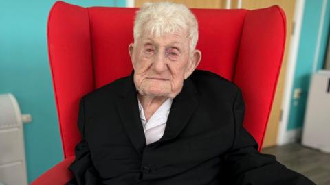 Donald Rose with short grey hair in a black blazer and white shirt sat in red armchair.
