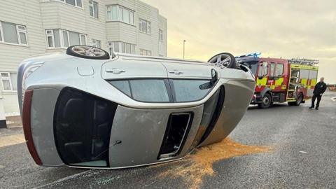 A silver, four-door car on its side on a road, with an open sunroof. A red fire engine can be seen in the background. There is a patch of sawdust around the car bonnet.
