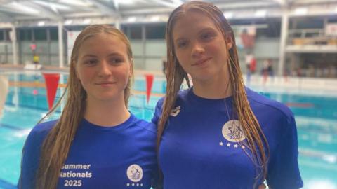 The sisters stand wearing blue tshirts in front a swimming pool. Their hair is damp and it looks like they've just finished a training session. 