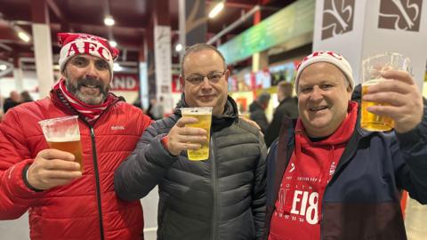 Three men, two of them wearing AFC hats raising a glass a beer inside a football stadium