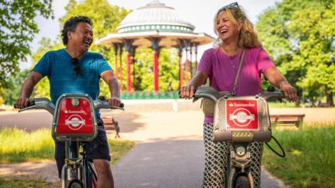 A man, in a blue top and shorts, and a woman, in black and white trousers and pink top, are cycling on red Santander rental bikes along a paved path in a sunny park. Behind them is a pavilion, surrounded by green trees and grass.