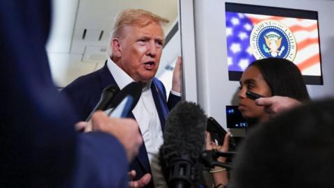 President Trump on Air Force One being interviewed by journalists. He is standing beside a video screen.