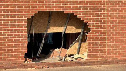 A large hole can be seen in a brick wall where a car smashed through it. Bricks and rubble can be seen inside the hole.