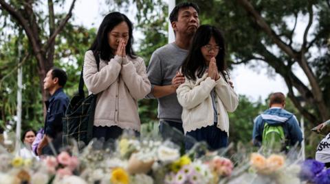 People praying as they stand in front of flowers laid on the ground as tribute to the victims of the fire