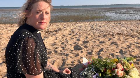 A woman with blonde curly and wavy hair kneels on a sandy beach looking at the camera. She wears black including a white dog collar. Behind her is the sea. Beside her is a bouquet of flowers.