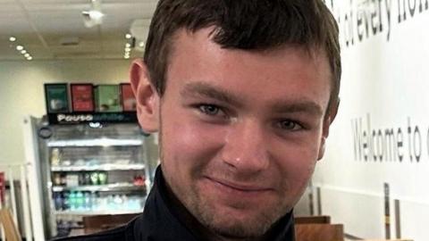 A man wearing a black round-necked top. He has short, brown hair and stubble and he is smiling.