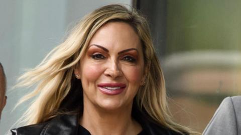 Jodie Marsh has long blonde hair and is wearing a black leather-style jacket as she exits a court building with a glass facade.