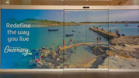A large poster covers a wall and a door. It features a sunny seaside scene with adults and children exploring rock pools beside a wall and standing on the wall further out into the the sea. Small boats are also in the sea.