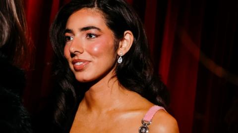 A close-up shot of Izzy Escobar standing against a red curtain. She looks to the side and wears sparkling silver earrings and has long black wavy hear tucked to one side behind her ear