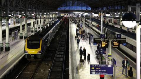 Manchester Picadilly station, platforms 6 and 7, as seen from above. A yellow and blue train is stopped at a platform and people are seen standing and sitting at the platform opposite. 