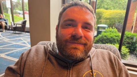 Mickey Bushell smiles at the camera, he is wearing a brown hoodie. In the background a woman is sitting in a chair