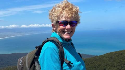 Liz Odell smiling at the camera while hiking up a mountain. She has short blonde curly hair and is wearing purple polarised mountaineering sunglasses, a blue half-zip fleece and a black back park.