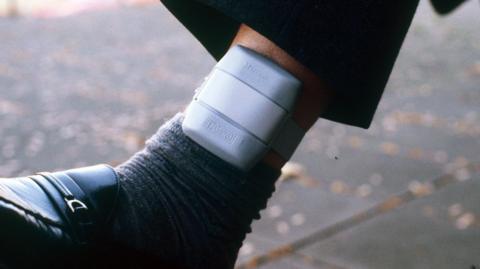 A close-up image of a grey ankle monitor. It is above a dark grey sock and black shoe on someone's leg 