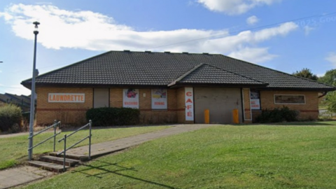 The brown boarded-up building has a triangular roof and there are steps leading down from it. The sky is blue and cloudy and there is a patch of grass in front of the building. There are also red-lettered signs on the building saying cafe and laundrette. 
