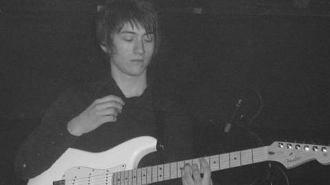 A young man with long dark hair playing a white guitar