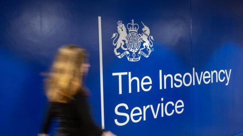 A blurred woman walking past a large blue wall which says The Insolvency Service under a crest in white lettering.