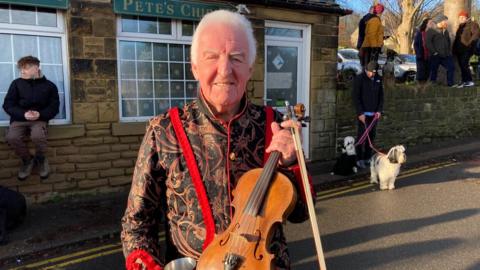 Ray Ellison stands outside the Old Harrow Inn, a traditional-looking stone-built pub, in Sheffield. He is holding a fiddle and bow and wearing a traditional sword-dancing jacket, which is black with an elaborate paisley design in shades of red and white.