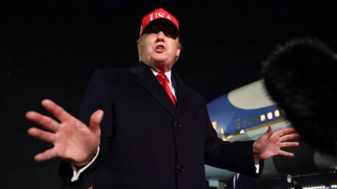 Donald Trump pictured from below with his hands outstretched, wearing a red USA baseball cap, with a plane visible in the background