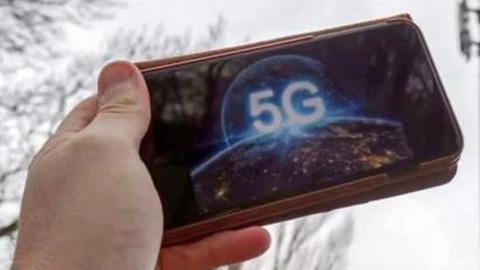 A hand holding a mobile phone with the letters 5G on it, with a mobile phone mast in the background