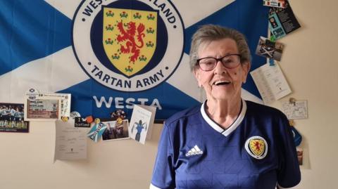 Moira, an elderly woman with short greyish hair and glasses, is standing on the left looking at the camera and smiling. She is wearing blue Scottish national football team jersey featuring team's crest.